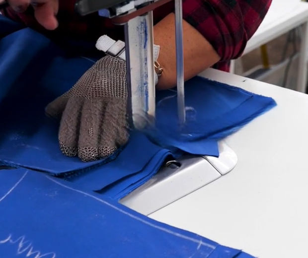 A hand wearing a protective glove guides a blue fabric under a sewing machine needle, highlighting textile work in progress.
