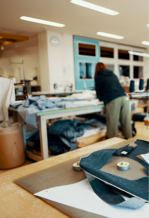 A tailor works at a cluttered workshop table with denim scraps and tools, background showing a bright, organized studio space.