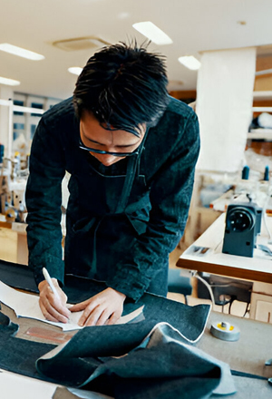 A person in a dark apron sketches patterns on fabric in a sewing workshop surrounded by sewing machines and tools.